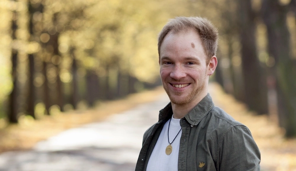 Ein lächelnder Mann mit einem verschwommenen Wald im Hintergrund.