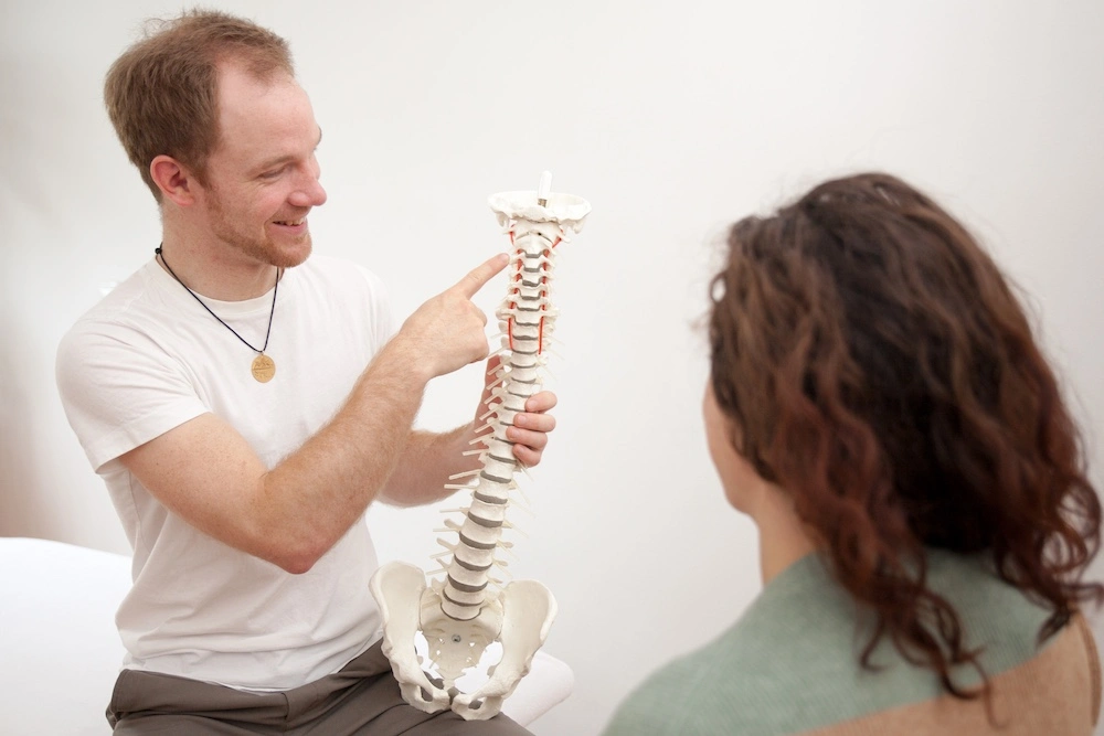 Ein Mann zeigt auf ein Modell einer Wirbelsäule. Eine Frau sitzt ihm gegenüber und hört ihm zu.